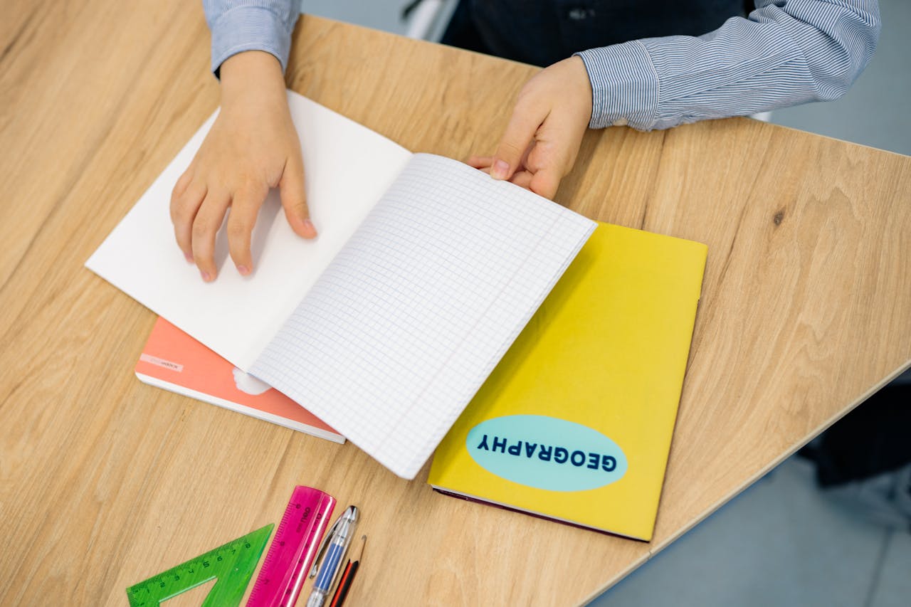 Hands flipping through a geography notebook on a wooden desk with pens and rulers.